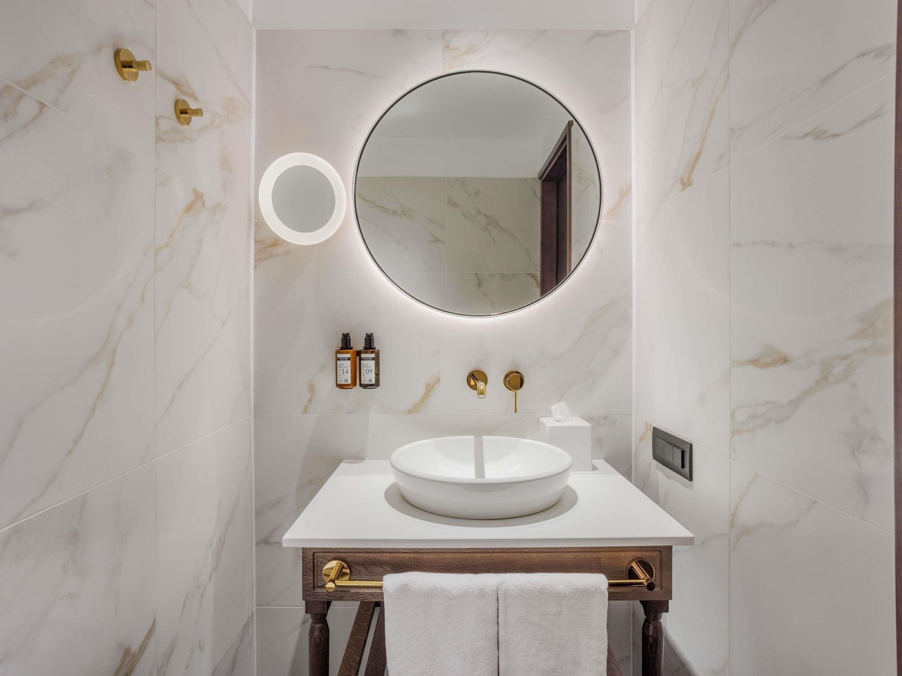 A bathroom with a round mirror, white sink, marble walls, and gold fixtures.