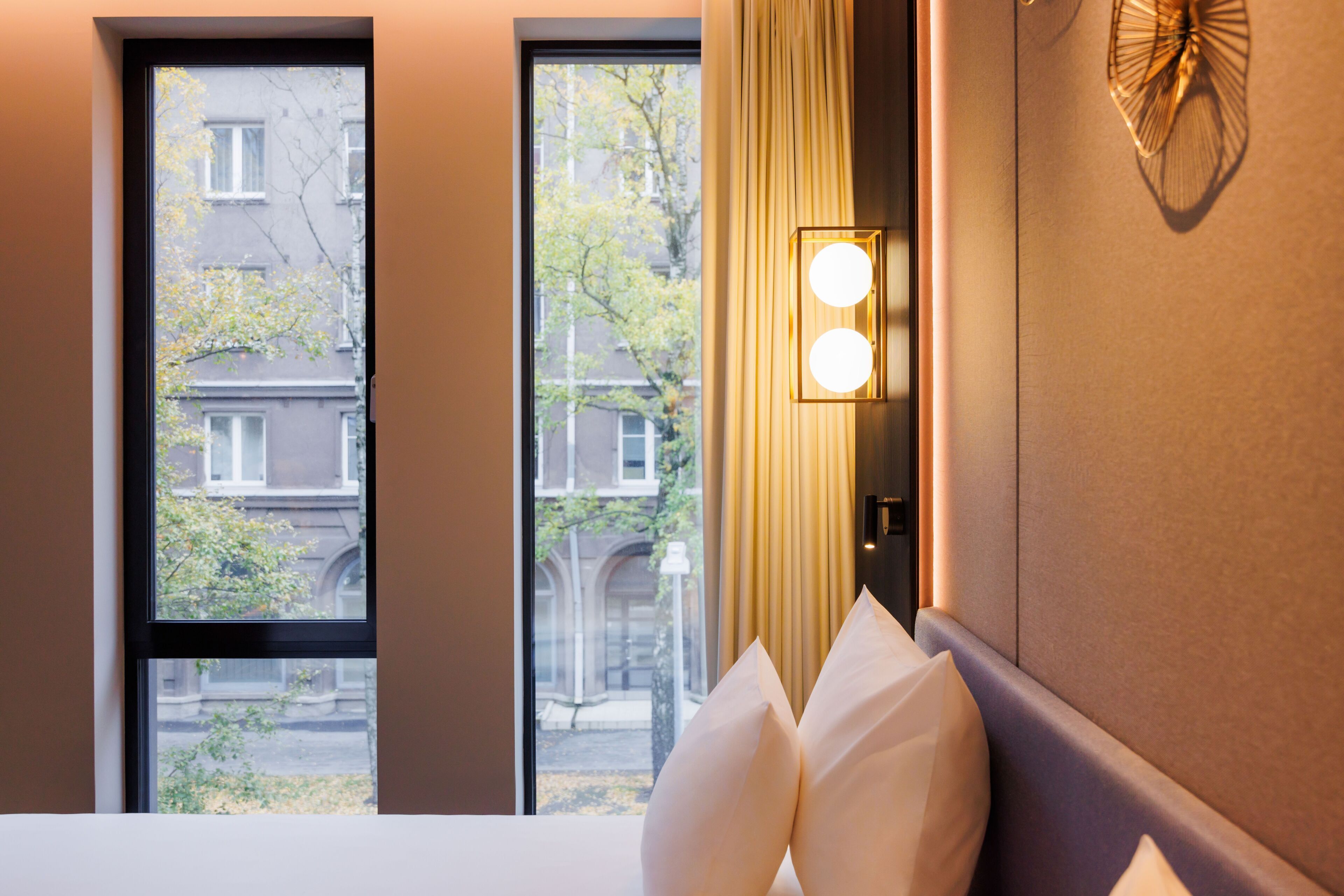 Een hotelkamer met een groot raam, een bed met witte kussens, een wandlamp en een decoratief kunstwerk.