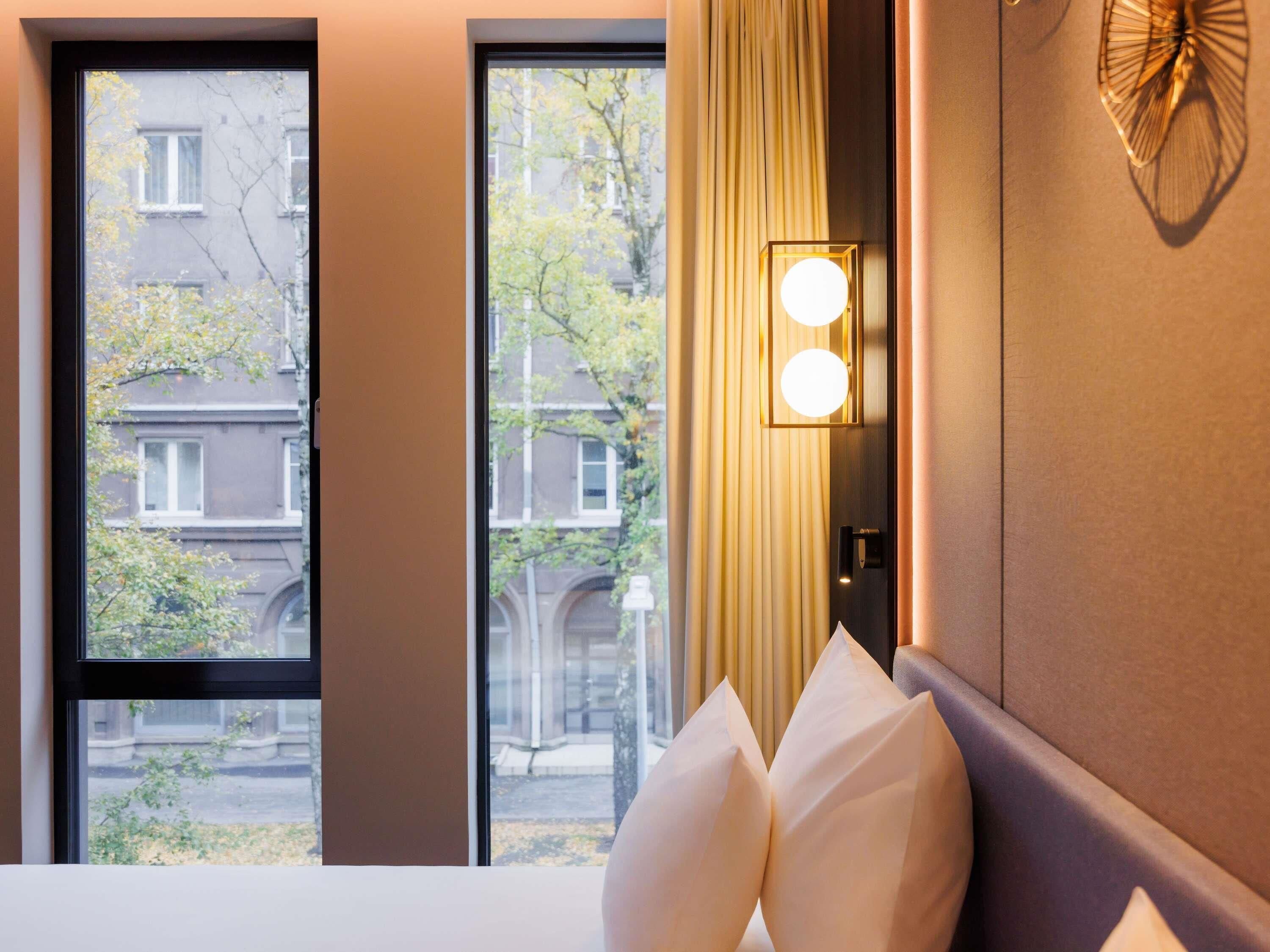 Habitación de hotel con un ventanal grande, una cama con almohadas blancas, una lámpara fijada a la pared y un adorno mural.