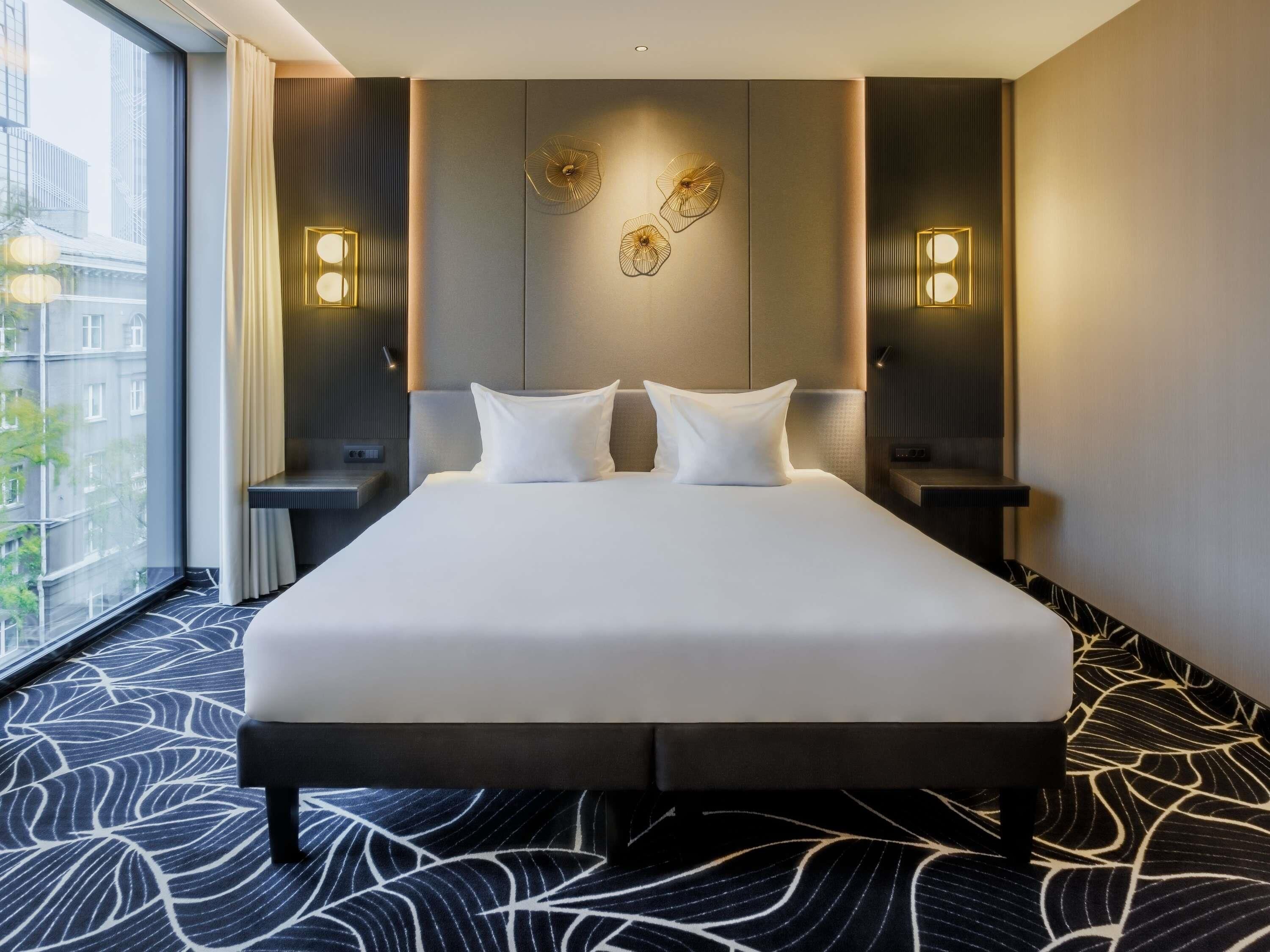 Quarto de hotel com uma cama grande, duas mesas de cabeceira, arandelas e vista para prédios por uma janela ampla.