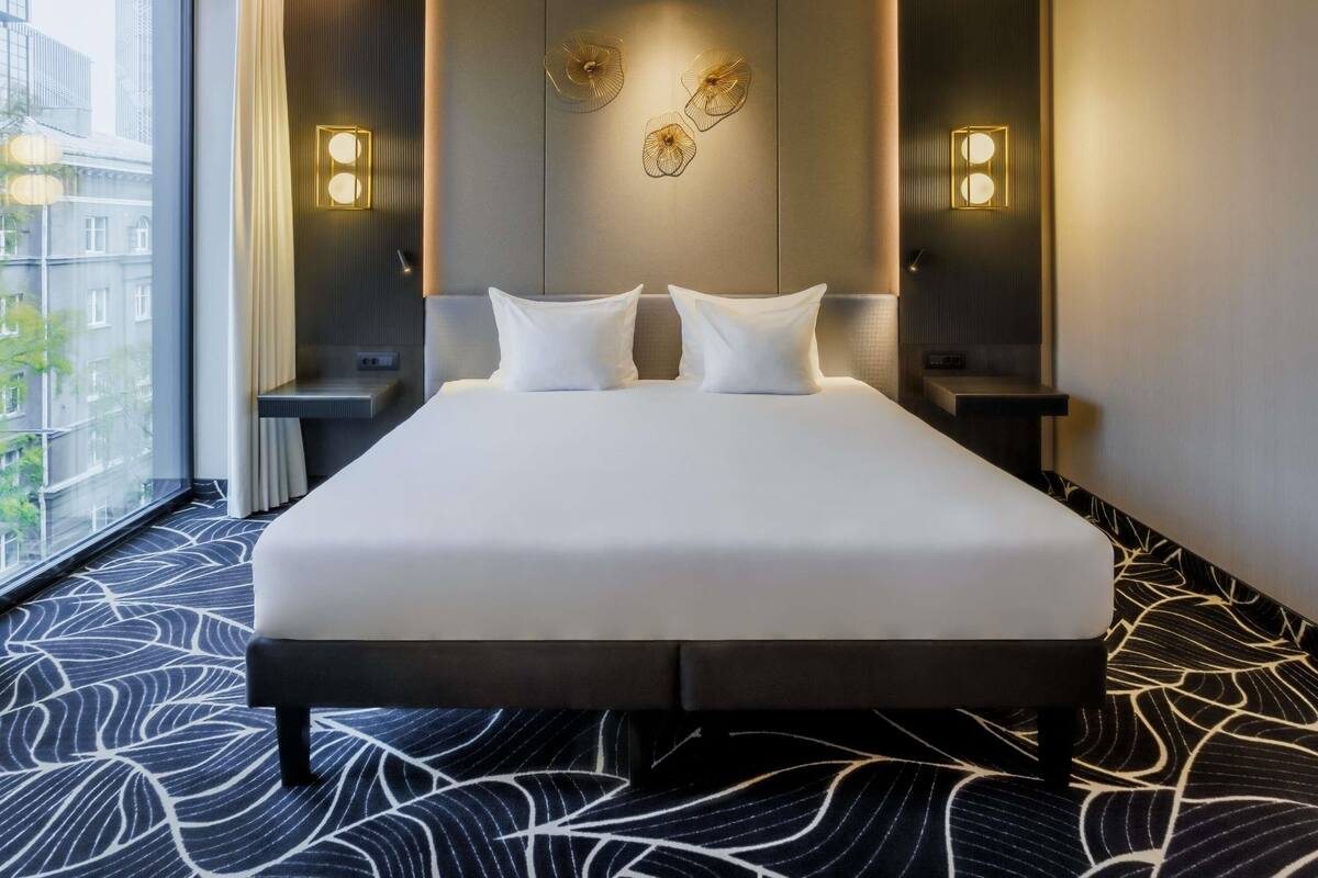 Ein Hotelzimmer mit einem großen Bett, zwei Nachttischen, Wandleuchten und Blick auf Gebäude durch ein großes Fenster.