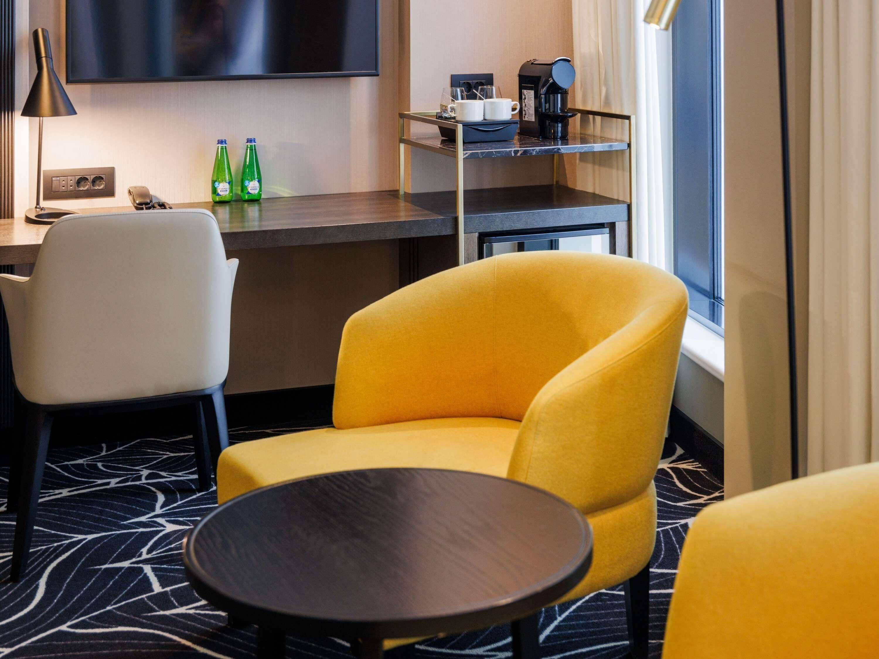 A hotel room with a yellow armchair, a white chair, a small round table, a TV mounted on the wall, a coffee machine, and a shelf with cups and bottles.