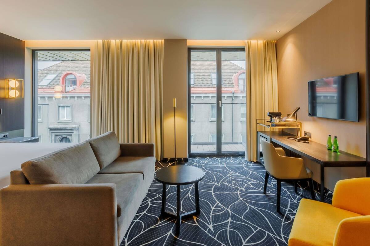 Ein Hotelzimmer mit Sofa, Schreibtisch, Stuhl, Fernseher und Blick auf Gebäude durch große Fenster.