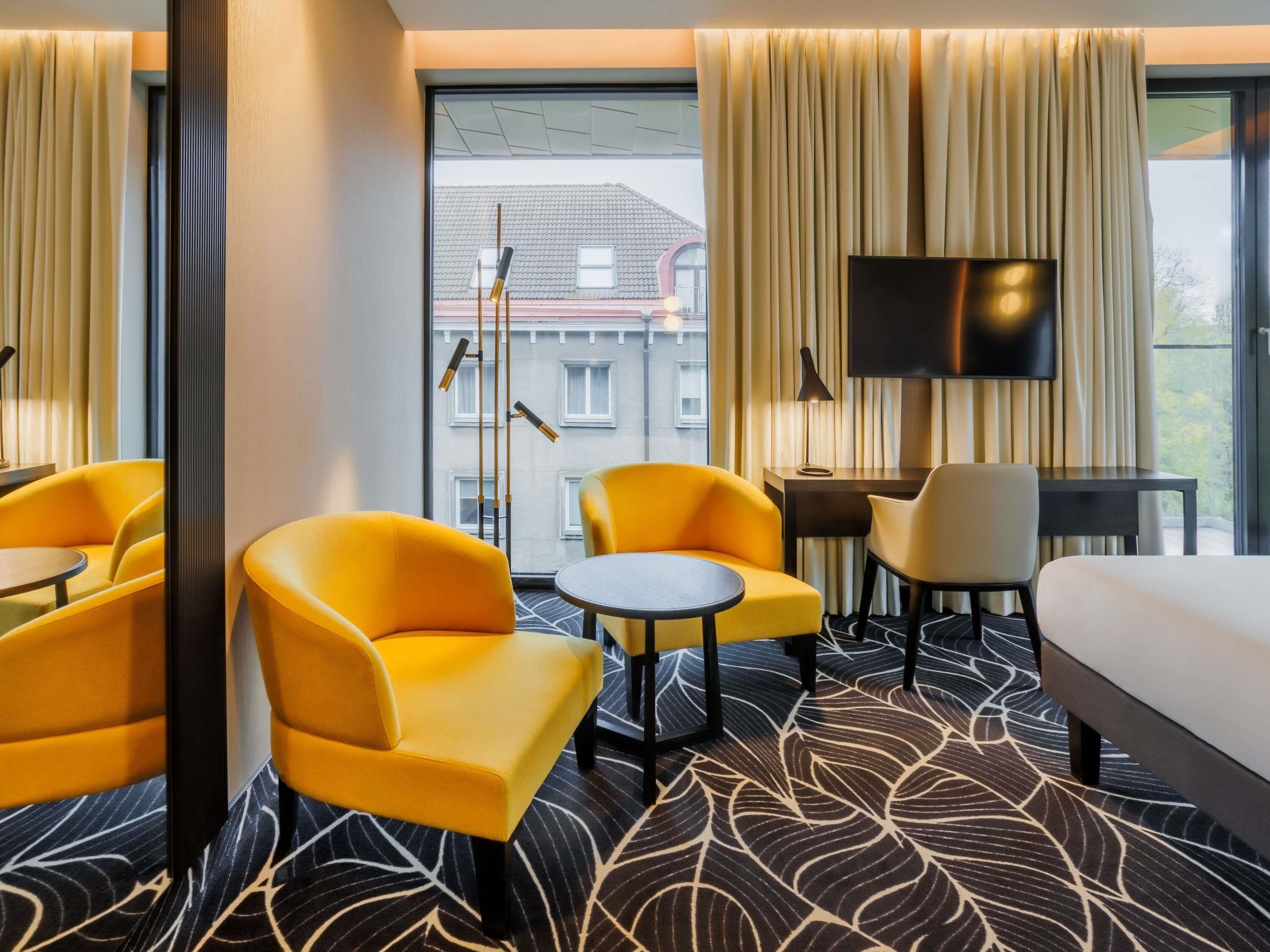 Une chambre d’hôtel avec un lit, deux fauteuils jaunes, une petite table ronde, un téléviseur et une vue sur les bâtiments environnants.