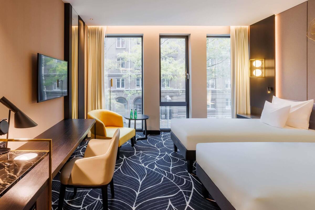 Ein Hotelzimmer mit Bett, Schreibtisch, Stuhl, Fernseher und Blick auf Gebäude durch große Fenster.