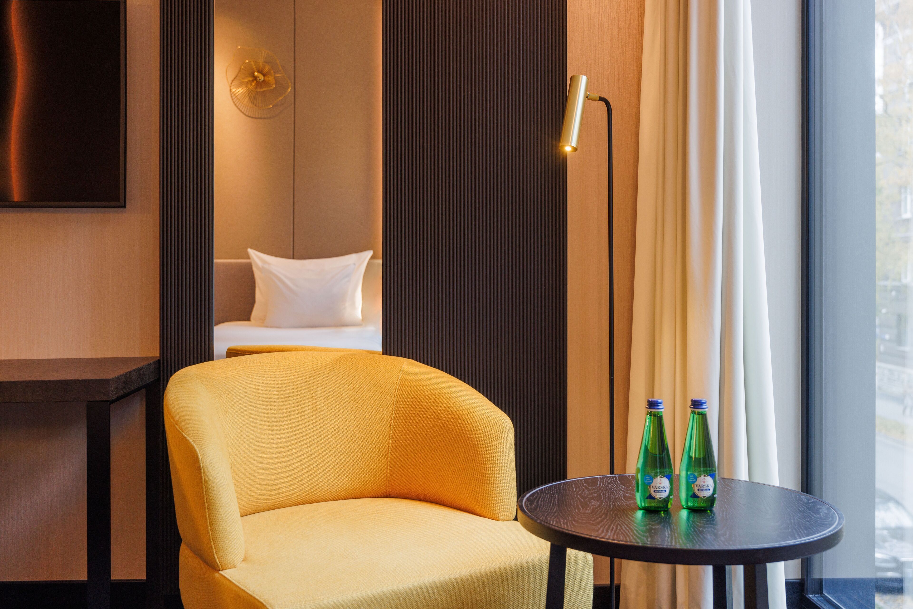 A hotel room with a yellow armchair, a small round table with two bottles, a bed with white bedding, a wall-mounted TV, and a window with curtains.