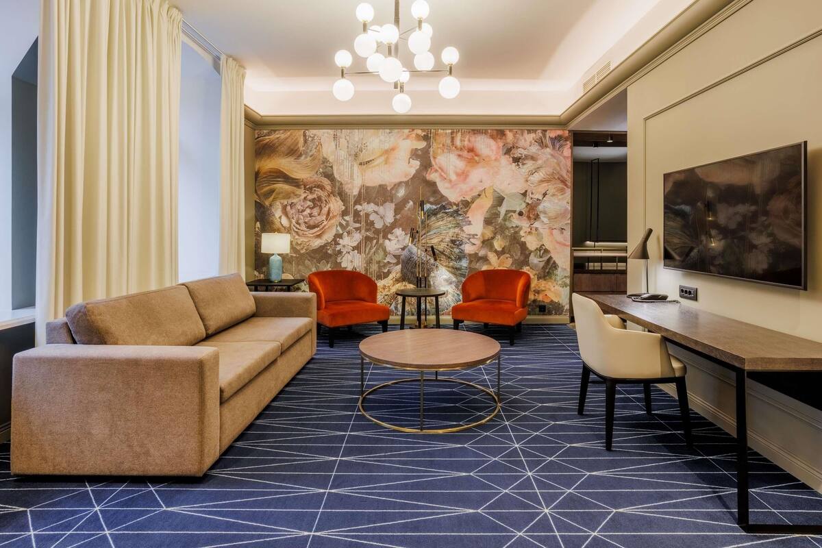 Ein Hotelzimmer mit einer Couch, zwei Sesseln, einem runden Couchtisch, einem Fernseher und einer Wand mit einem floralen Wandbild.