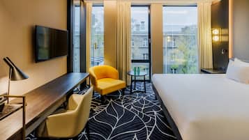 Habitación de hotel con una cama grande, un escritorio con una silla, una mesita con otra silla y un televisor montado en la pared.