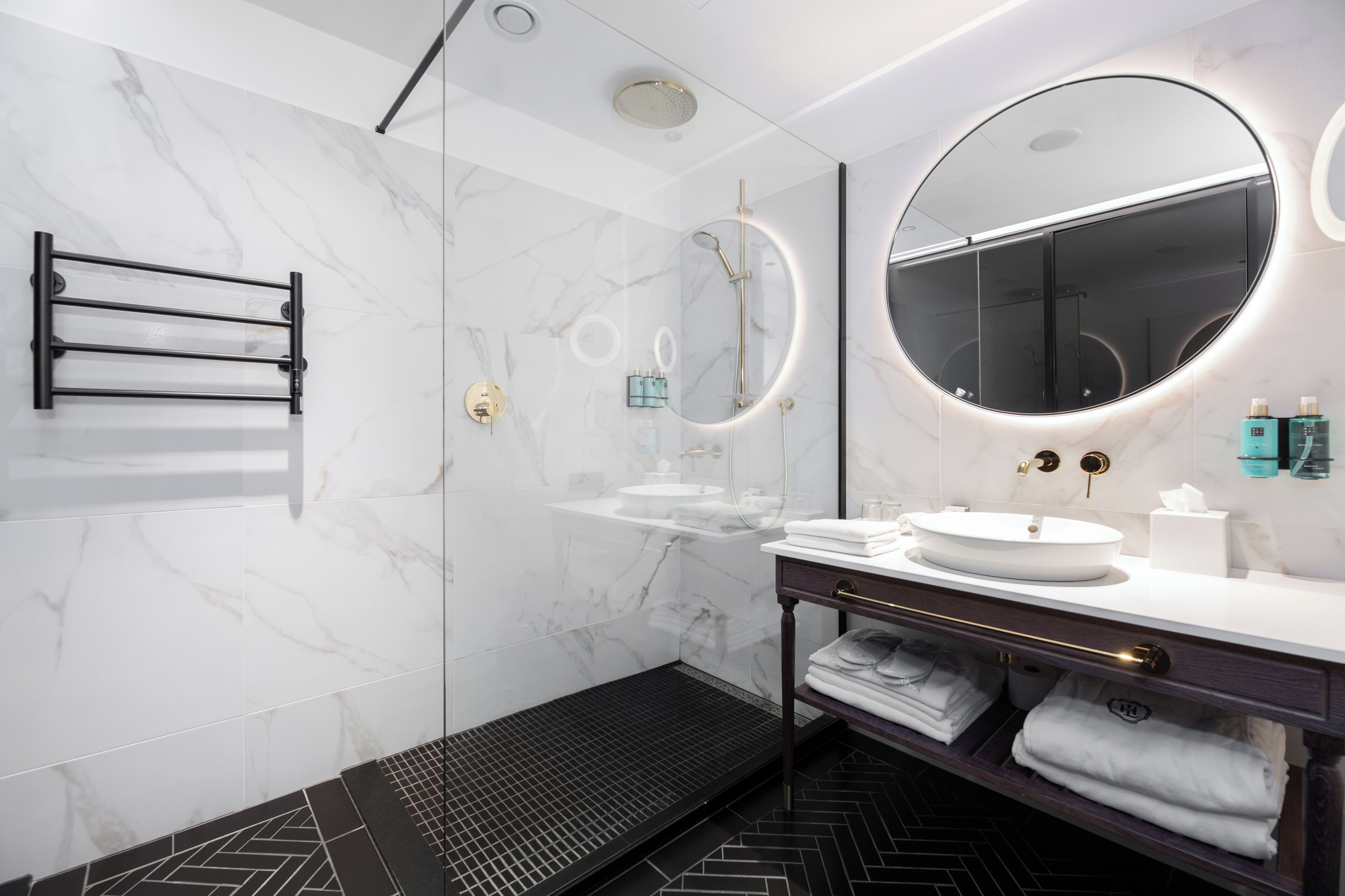 A modern bathroom with a glass-enclosed shower, a round mirror, and a vanity with a sink and towel rack.