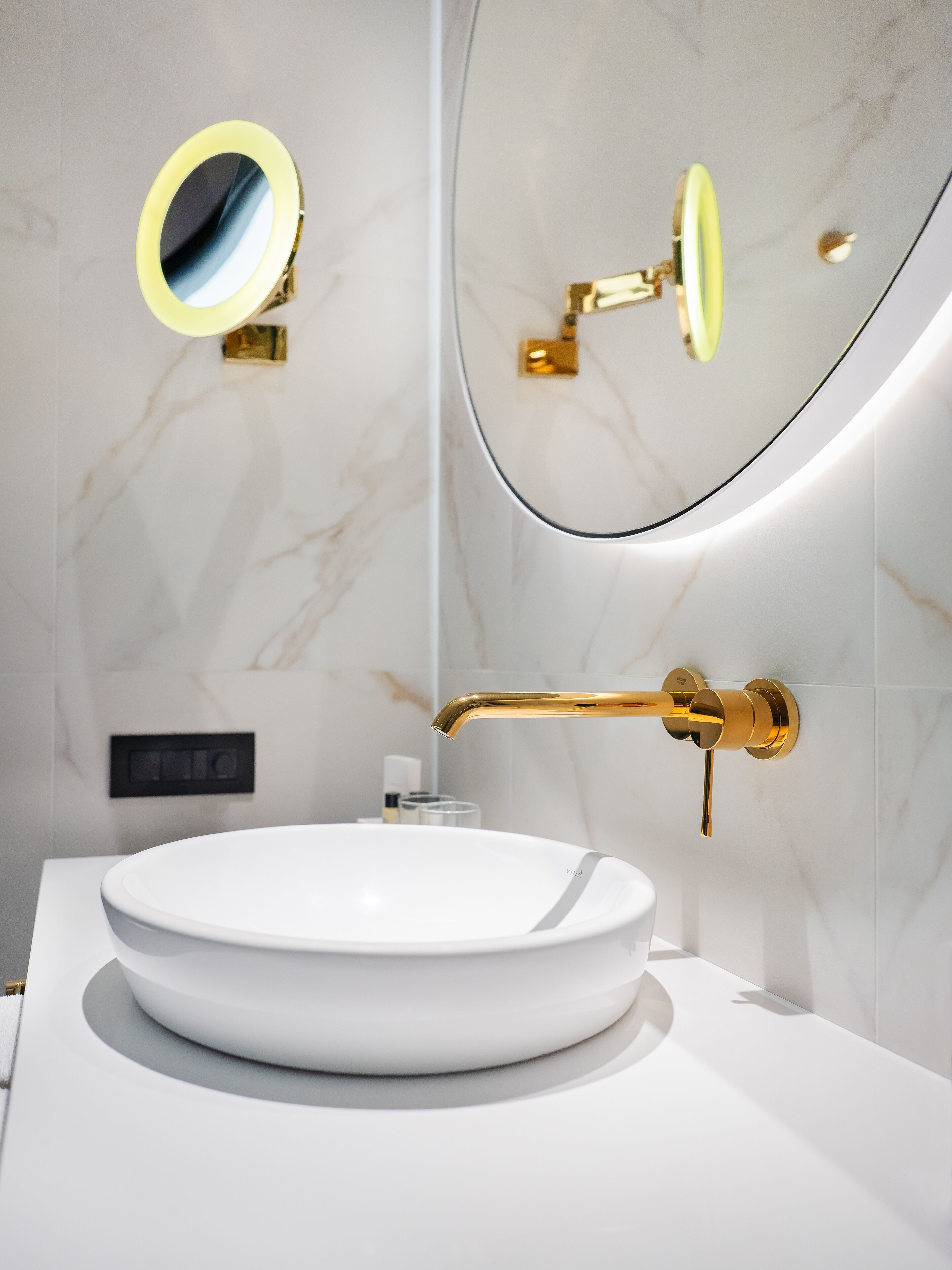 A modern bathroom with a round white sink, gold fixtures, and a marble wall.