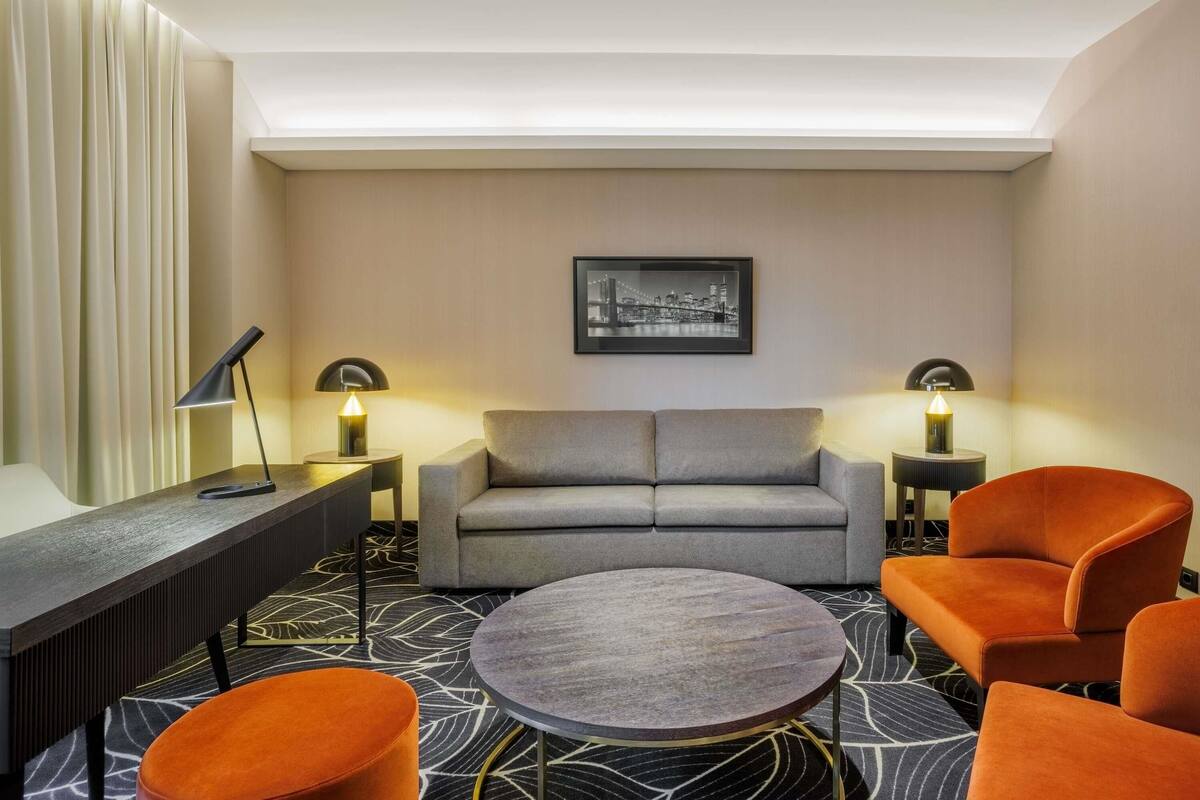 Una habitación de hotel moderna con un sofá gris, sillones naranjas, una mesa de centro redonda y dos lámparas de noche.