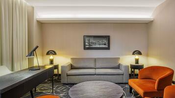 A modern hotel room with a grey sofa, orange armchairs, a round coffee table, and two bedside lamps.