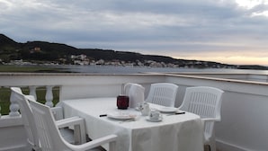 A table set for two with white chairs and a tablecloth, overlooking a body of water and a hillside with buildings. - Classic Attic Apartment (Rab)