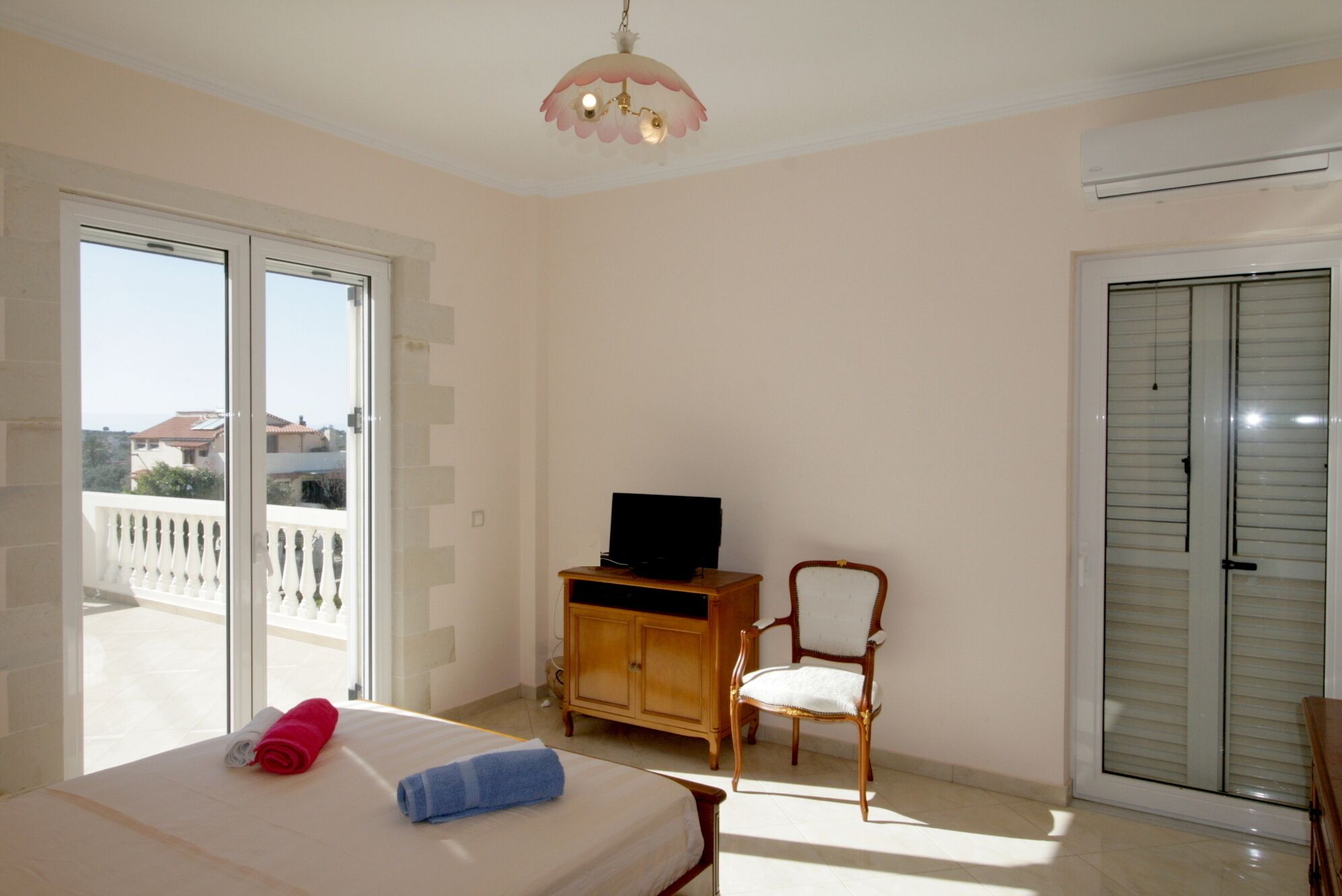 Un dormitorio con cama, televisor sobre un mueble de madera, una silla y un balcón con barandilla.