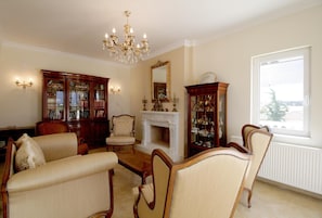 Ein traditionell eingerichtetes Wohnzimmer mit Kamin, zwei Sesseln, einer Holzvitrine mit Glastüren, einer Chandelierleuchtre und einem großen Spiegel.