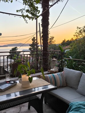 Un balcón con vista al mar, una mesa con una copa de vino tinto y un sofá de mimbre.