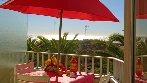 Ein Balkon mit einem roten Sonnenschirm, einem Tisch mit Früchten und einer Flasche, und Blick auf das Meer und Palmen.