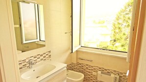Ein Badezimmer mit einem großen Waschbecken, einem Spiegel, einem Bidet und einem Fenster mit Blick auf Grünfläche.