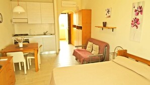 Ein kompakter Raum mit Bett, kleinem Schreibtisch, Stuhl, einer Küchenzeile und einer Tür zu einem weiteren Zimmer.