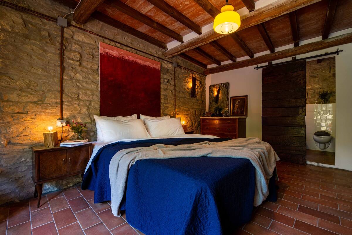 Un dormitorio con pared de piedra, vigas de madera, cama con ropa de cama azul, una mesita de noche y una pared de acento roja.