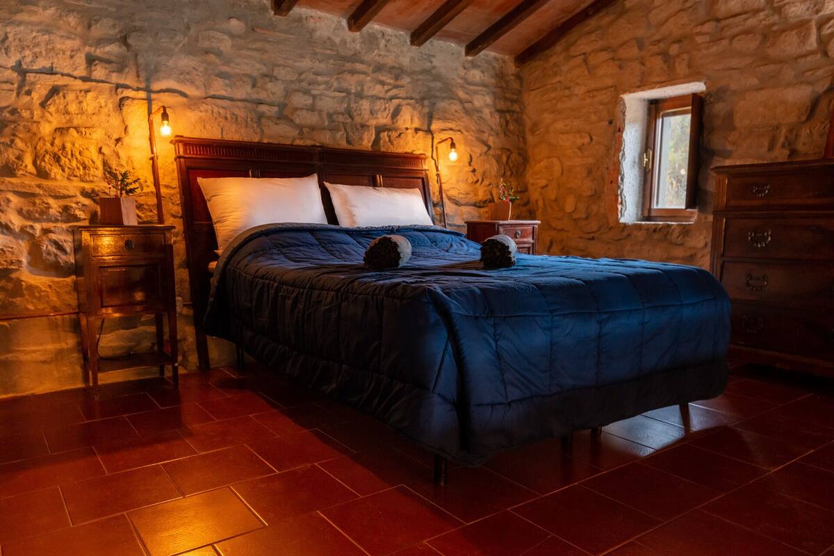 Un dormitorio con pared de piedra, cama de madera con colcha azul oscuro, dos mesitas de noche con lámparas y un aparador de madera.