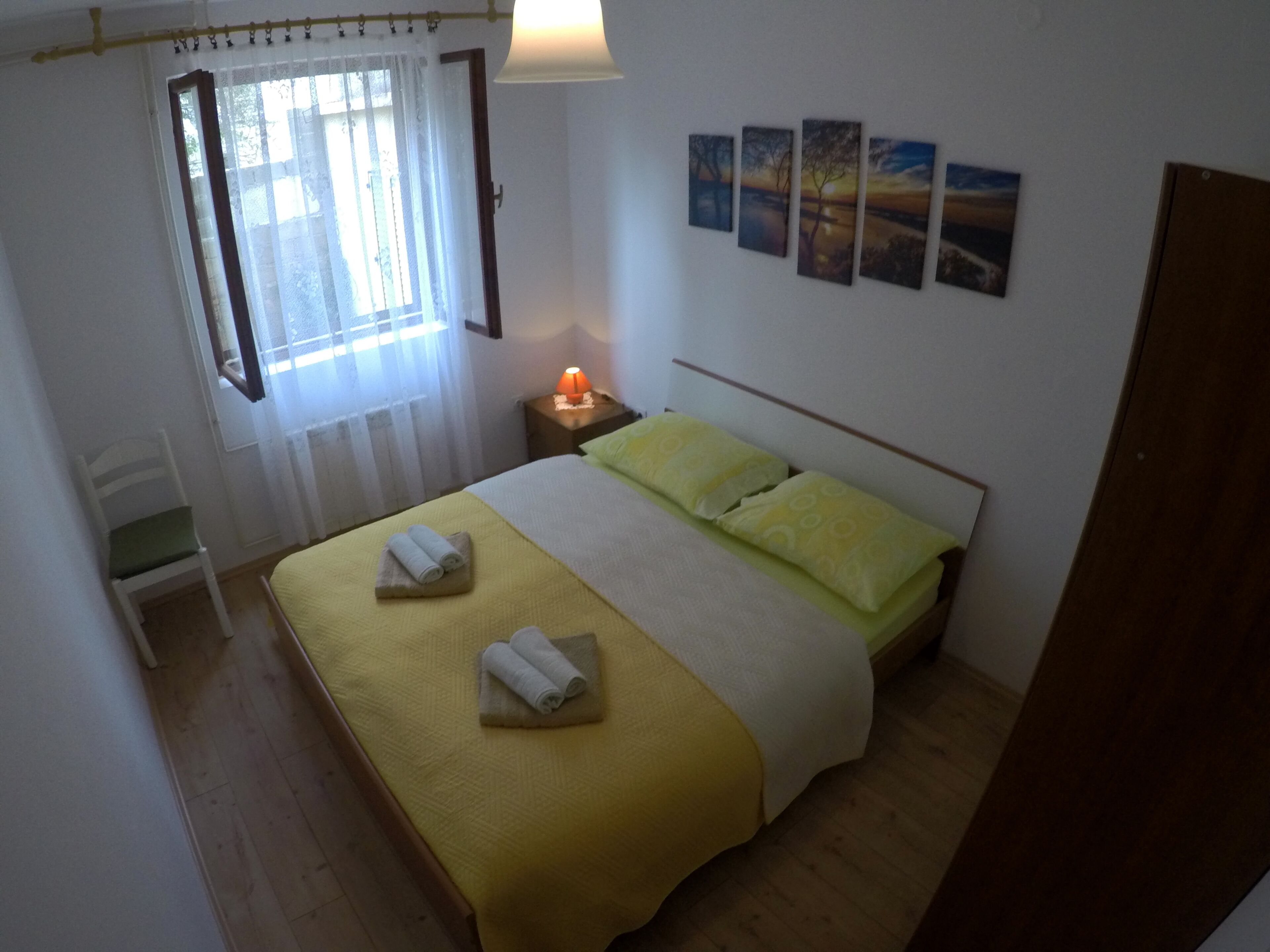 Een slaapkamer met een bed, nachtkastje, lamp, stoel en een raam met gordijnen.