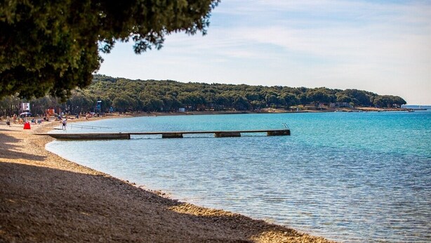 Une plage avec une jetée en bois qui s’avance dans des eaux calmes, entourée d’une colline boisée et surplombée d’un ciel dégagé.