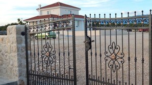 Une entrée fermée par un portail en fer forgé décoratif, un mur de pierre et un bâtiment avec un toit rouge en arrière-plan.