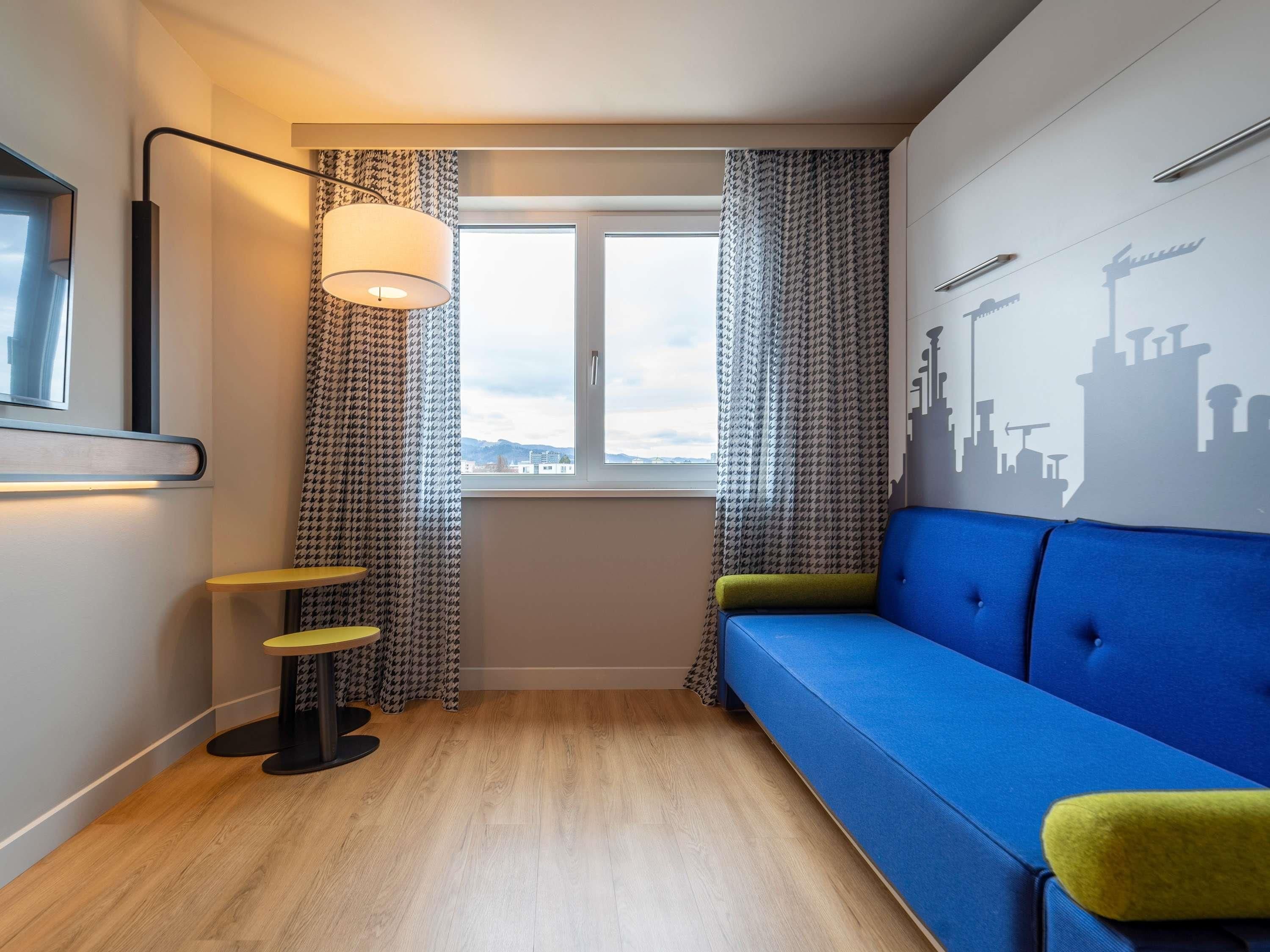 Una habitación con sofá azul, una mesita, una lámpara, cortinas estampadas y un mural de paisaje urbano.