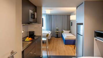A compact hotel room with a kitchenette, a bed, a small table with a lamp, and a window with patterned curtains.