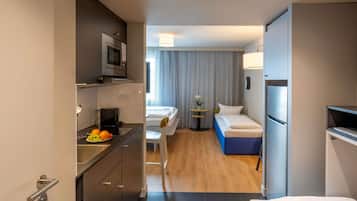 A compact hotel room with a kitchenette, a bed, a small table with a lamp, and a window with patterned curtains.