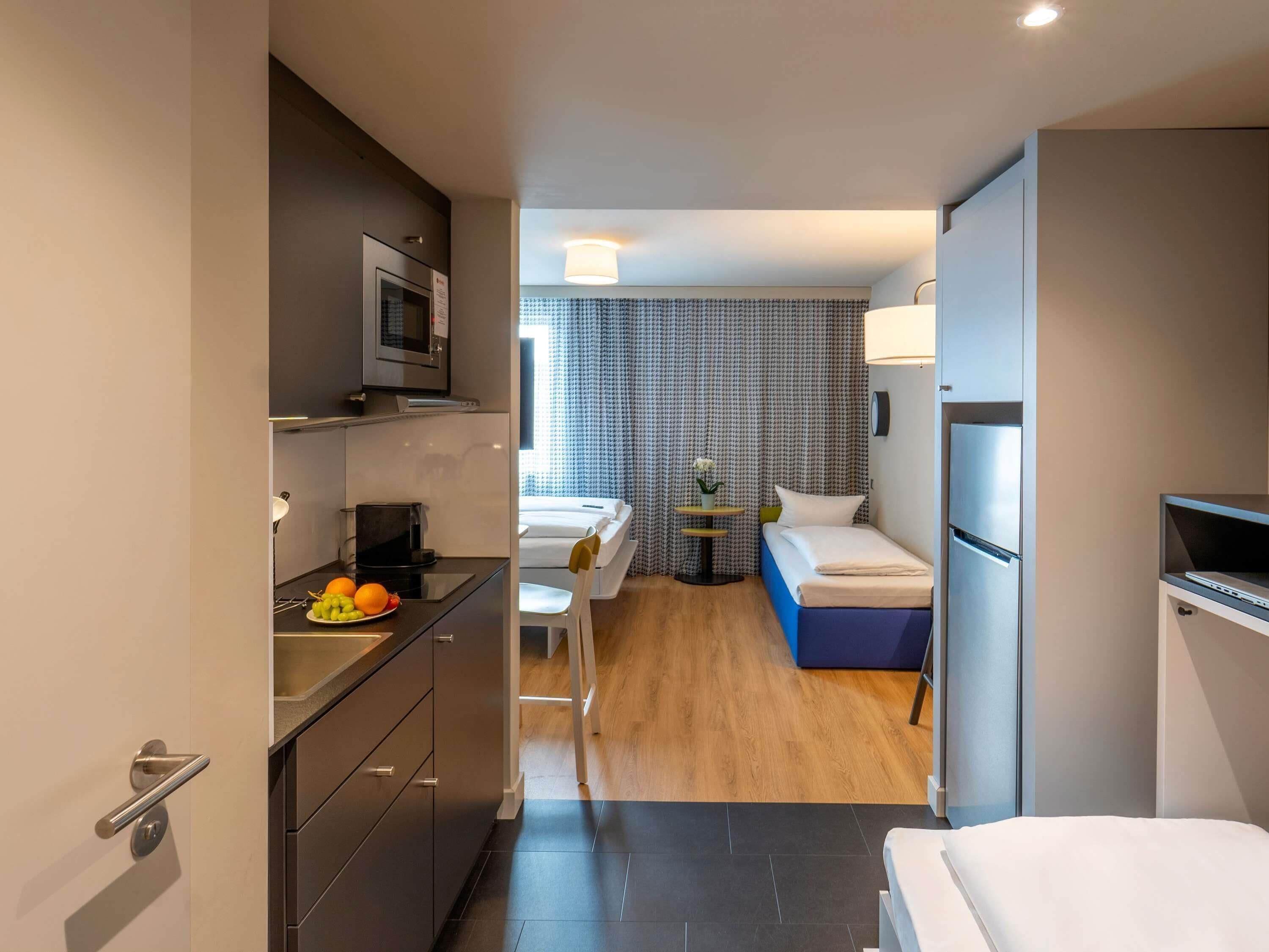 Een kleine hotelkamer met een kitchenette, een bed, een tafeltje met een lamp en een raam met gordijnen met een patroon.