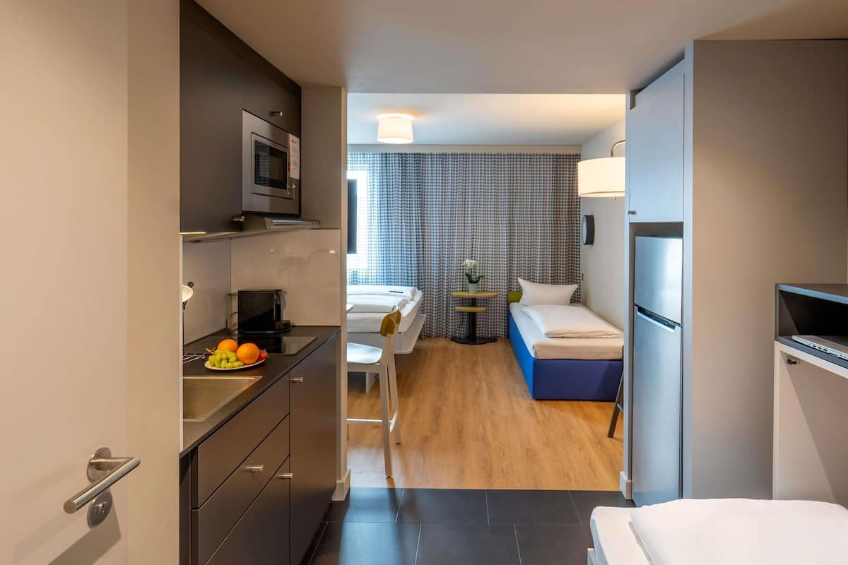 Een kleine hotelkamer met een kitchenette, een bed, een tafeltje met een lamp en een raam met gordijnen met een patroon.