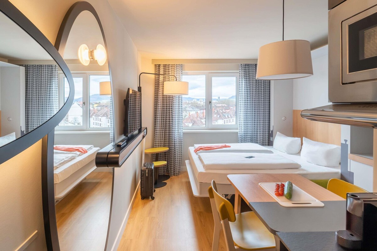 Une chambre d’hôtel comprenant un lit, un bureau avec une chaise, une télévision, un miroir et une fenêtre donnant sur des immeubles.