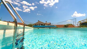 A clear swimming pool with a metal ladder on the left, surrounded by a fence. In the background, there is a building with a tiled roof and a clear blue sky with clouds. - Luxurious Villa in Ružići Croatia With Private Pool and Garden (Matulji)