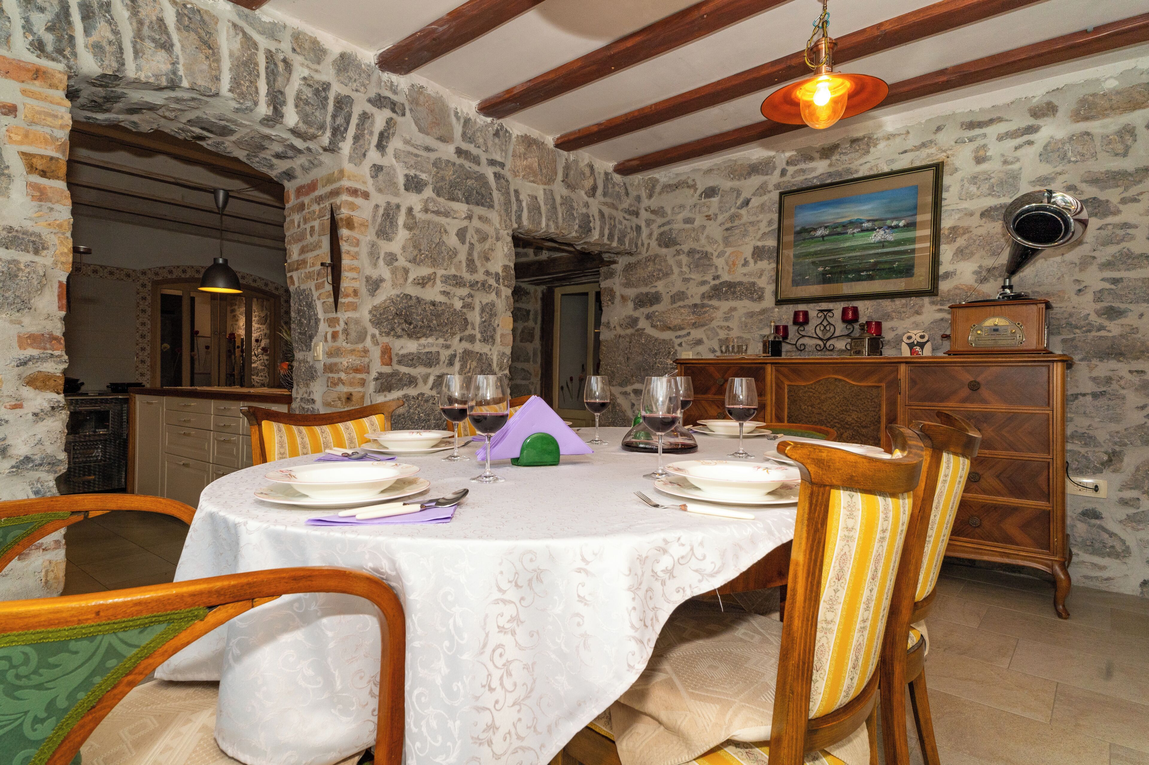A dining area with a round table set for six, surrounded by wooden chairs with striped cushions. A stone wall and wooden beams are visible in the background.