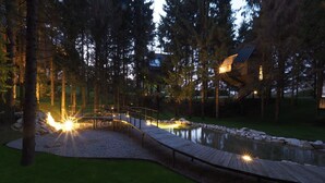 Eine Holzterrasse mit Geländer, ein kleiner Teich und ein Gebäude mit geneigtem Dach in einem bewaldeten Gebiet.