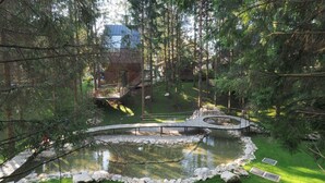 Ein bewaldetes Gebiet mit einem Teich, einer Holzbrücke und einem Gebäude mit einem Metall-Dach im Hintergrund.