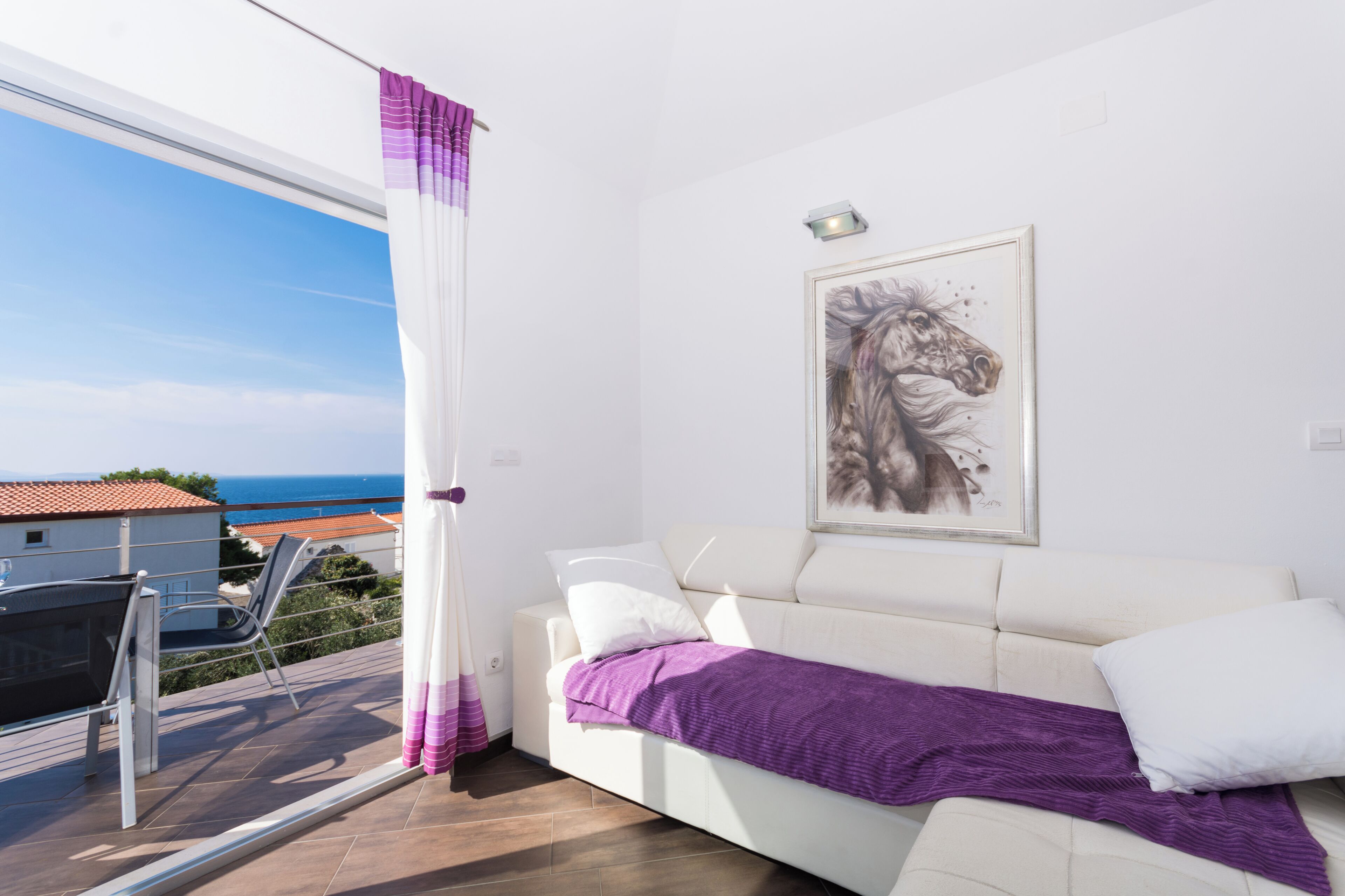 A white sofa with a purple blanket, a white pillow, and a framed artwork of a horse on the wall.