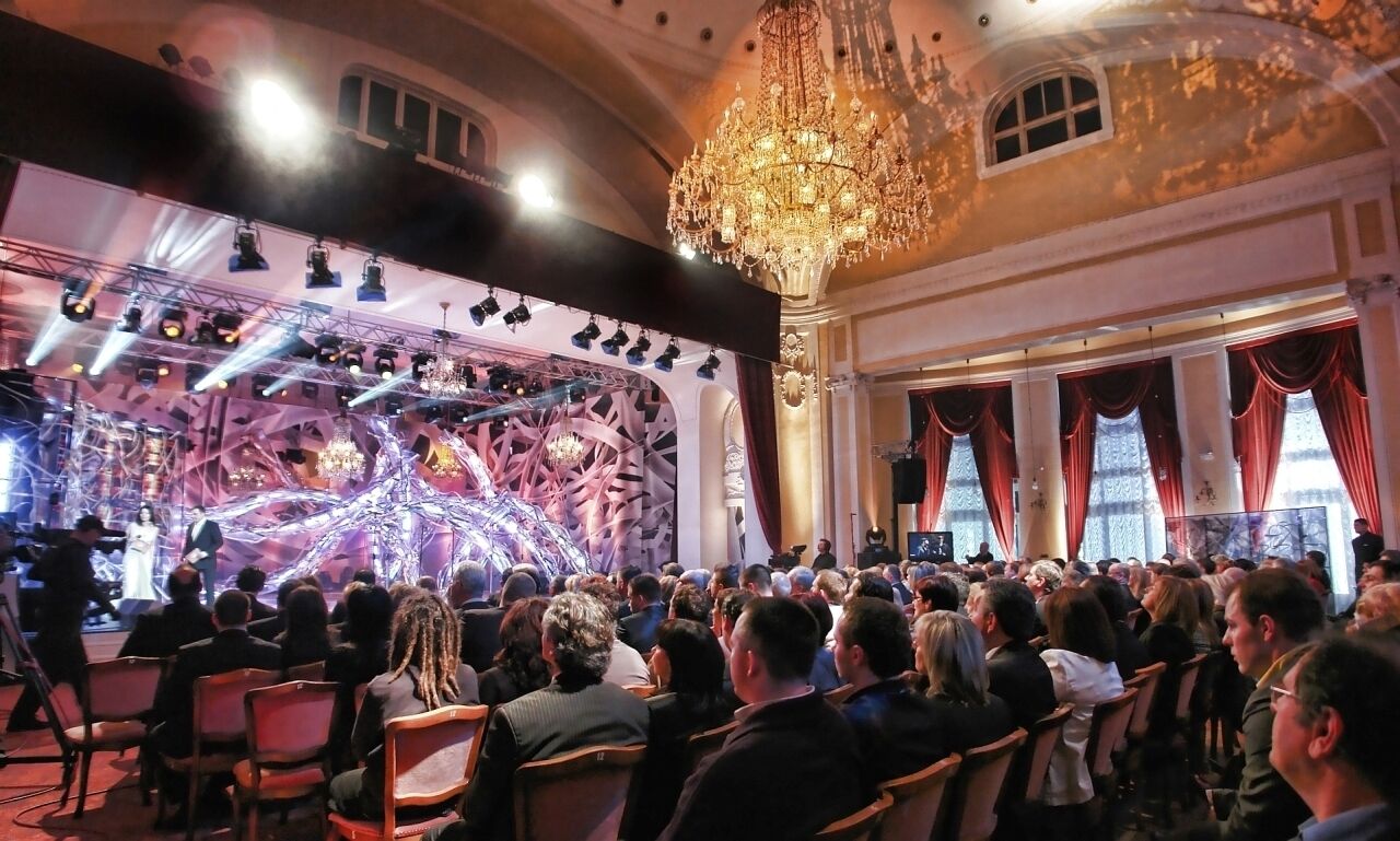 An indoor event with a stage, audience, and ornate chandelier.