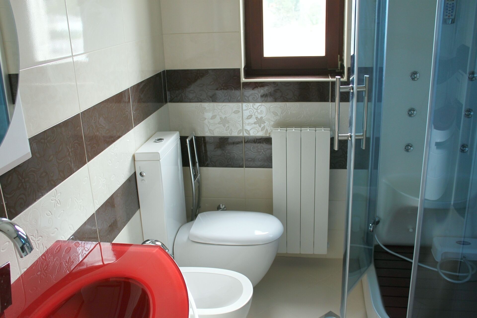 Une salle de bain avec des toilettes, un lavabo rouge, une douche en verre et des carreaux décorés.