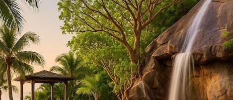 A pool with a waterfall, surrounded by palm trees and a gazebo.