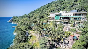 A coastal property with a large building featuring multiple balconies and a swimming pool, surrounded by lush greenery and overlooking the sea.