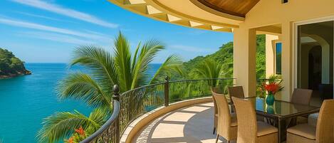 A balcony with a curved railing, a dining table with chairs, and a view of the ocean and greenery.
