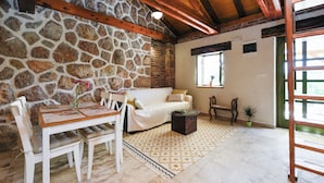 Un mur en pierre, des poutres en bois au plafond, un canapé blanc, une table à manger en bois avec des chaises, une table basse et un tapis à motifs.