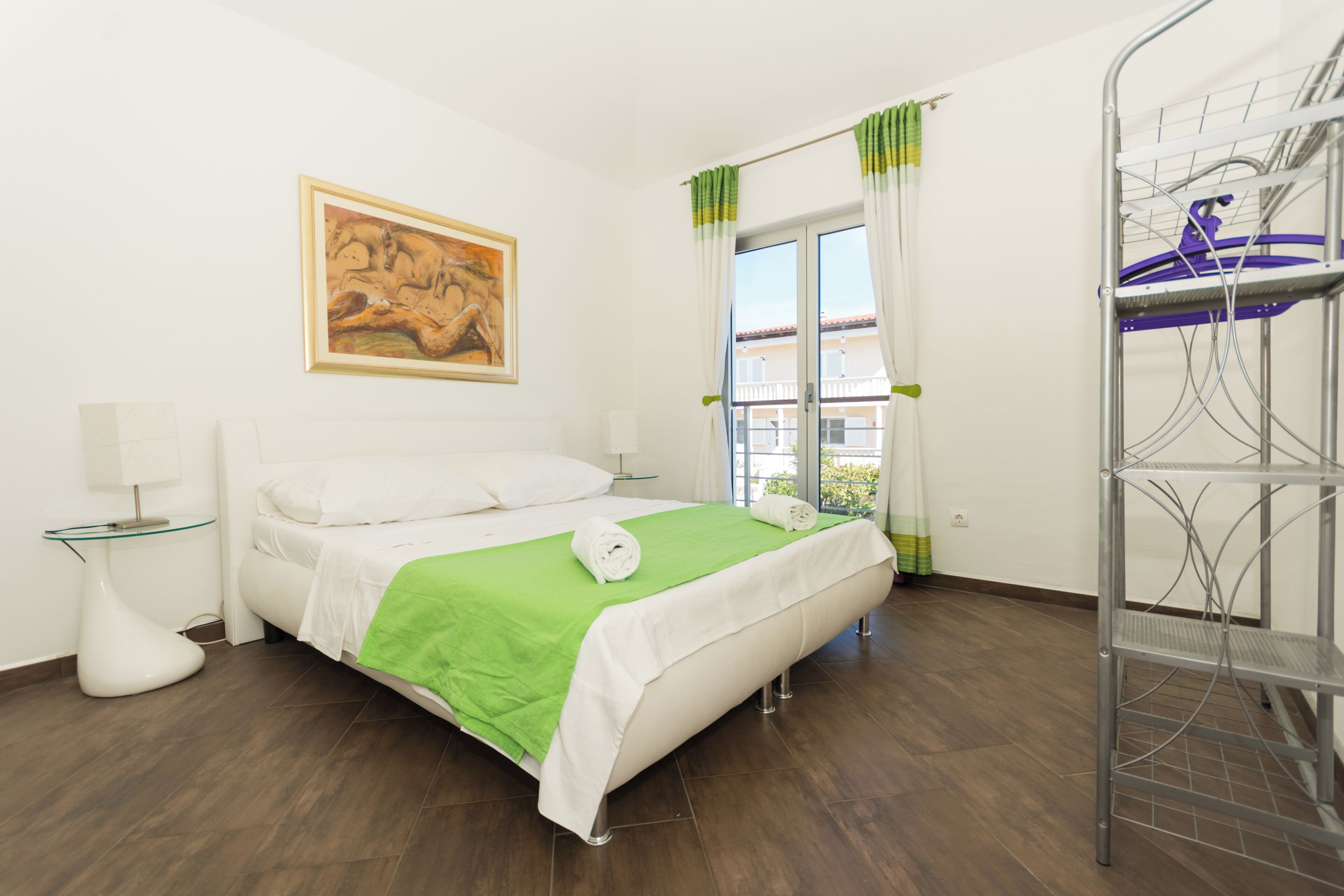 A hotel room with a bed, a painting on the wall, a window with curtains, and a metal shelf unit.