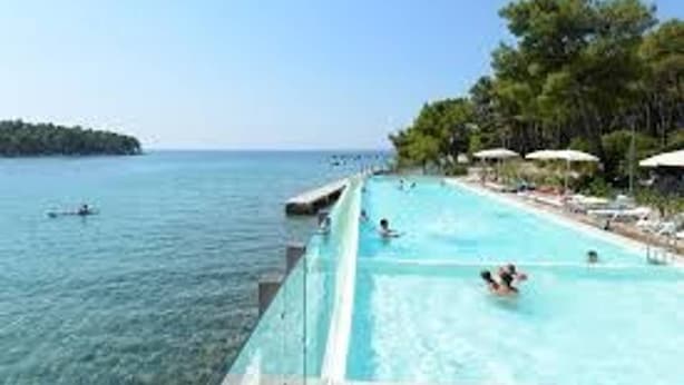 A swimming pool with clear blue water, a view of the sea, and a row of umbrellas along the poolside. - Room in Apartment - Great Location Just a Short Walk From the Beach and Downtown Biograd - 4 Guests! (Biograd na Moru)