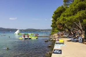 A beach with people swimming and boats in the water, a crowd on the sand, and trees in the background.