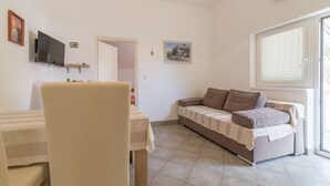 A living room with a sofa, a TV mounted on the wall, a table, and a window with blinds. - Room in Apartment - Great Location Just a Short Walk From the Beach and Downtown Biograd - 4 Guests! (Biograd na Moru)