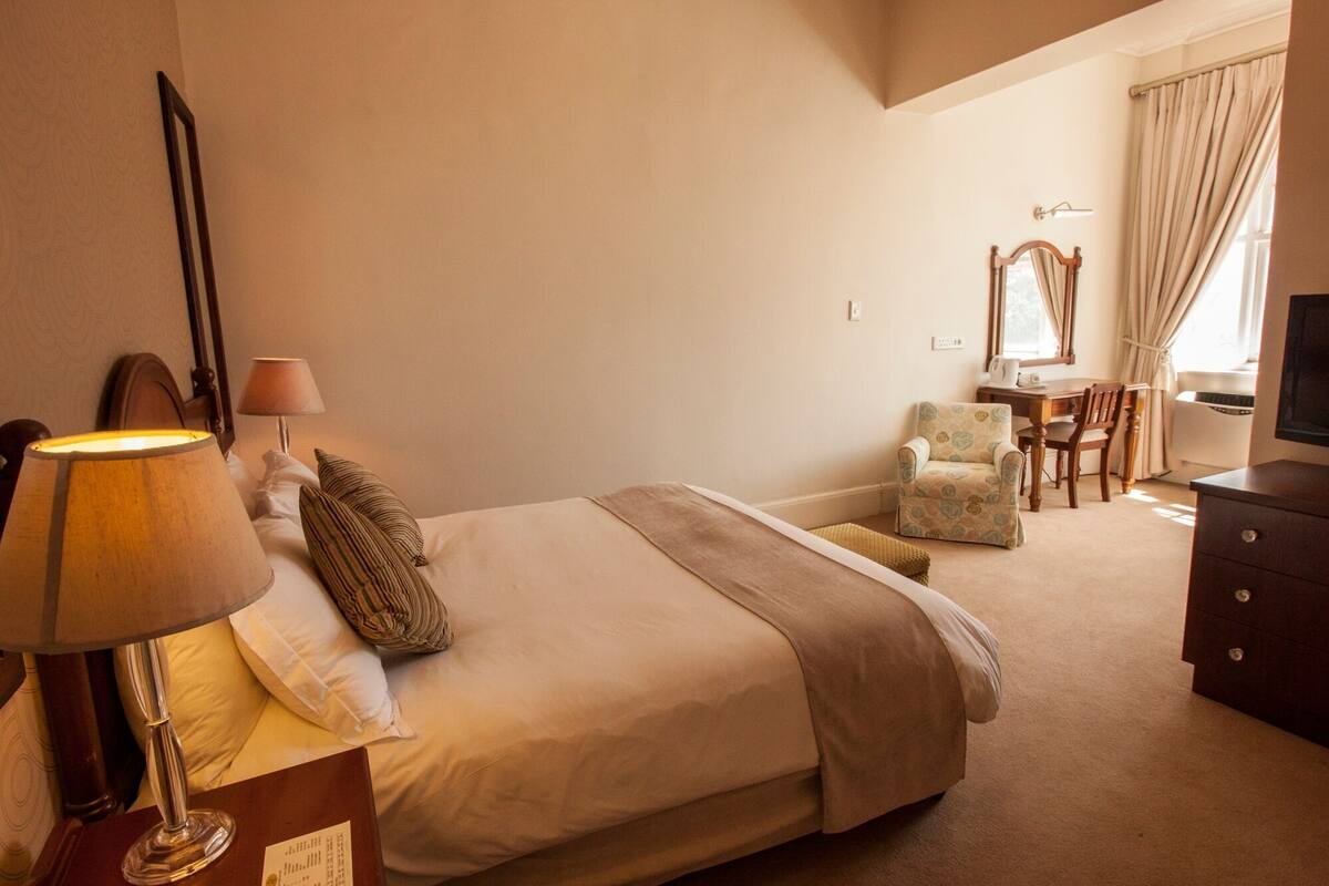 Ein Hotelzimmer mit Bett, Schreibtisch, Stuhl, Lampe, Spiegel und Fernseher.